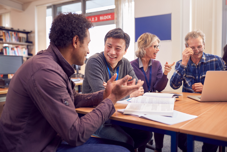 Teacher With Group Of Mature Adult Students In Class Sit Around Table And Work In College Library